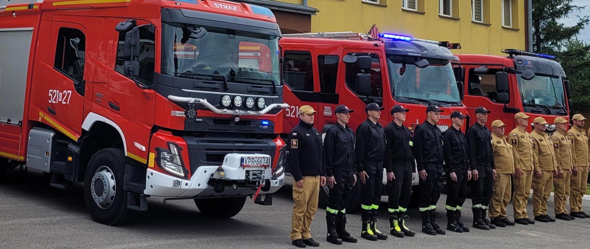 Strażacy stoją w szeregu przed samochodami pożarniczymi