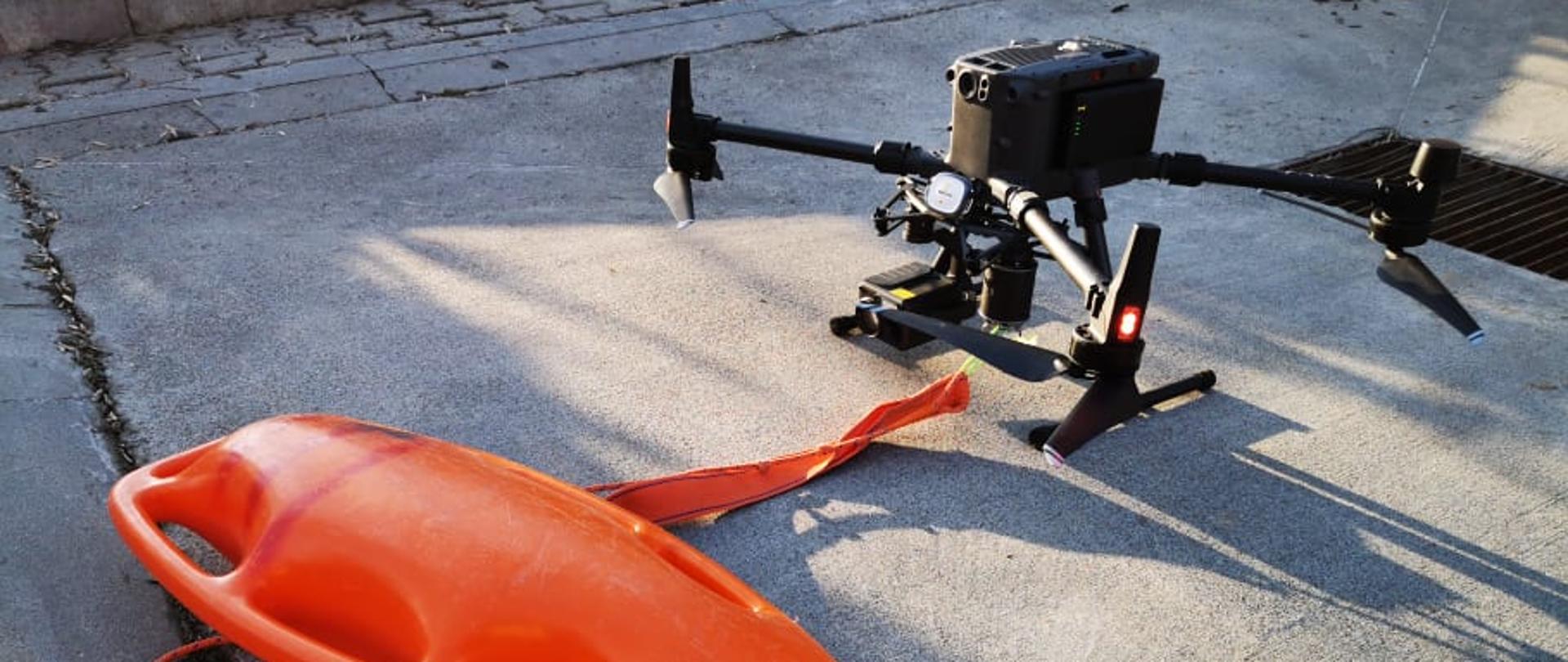 Na zdjęciu widoczny bezzałogowy wielowirnikowy statek powietrzny (dron), do którego linką przywiązana jest plastikowa boja ratownicza używana przez ratowników do podawania jej tonącym. Zestaw stoi na betonowym placu i jest przygotowywany do lotu.
