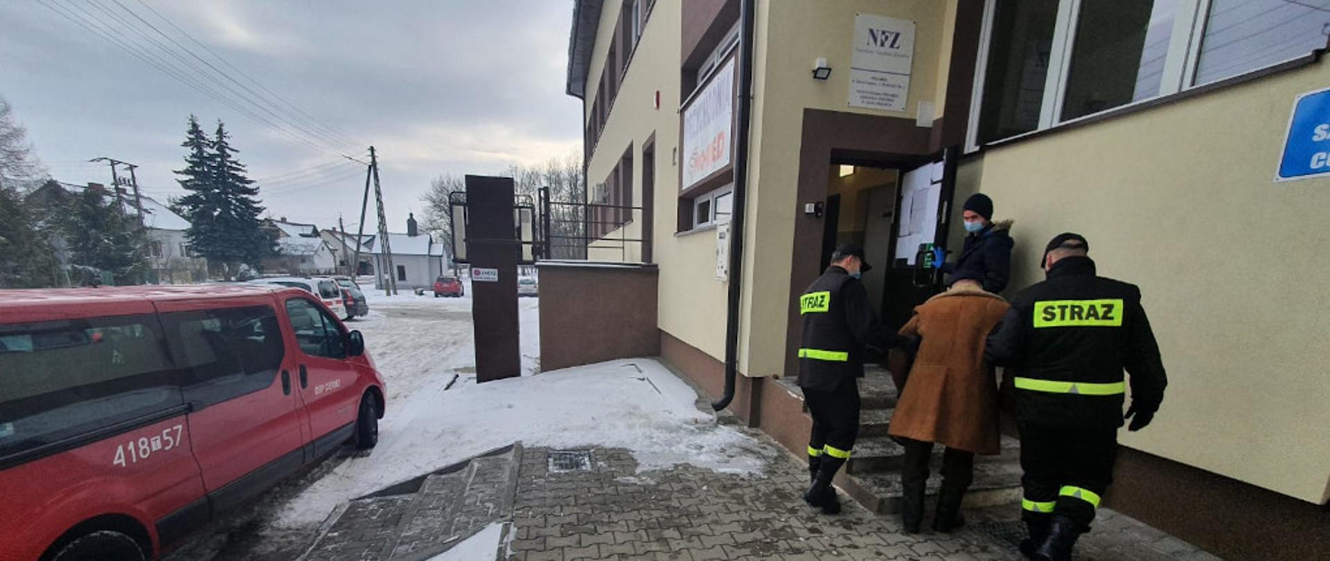 Na zdjęciu przedstawiony jest po lewej stronie samochód operacyjny OSP. Po prawej stronie ukazani są dwaj strażacy w umundurowaniu koszarowym z założonymi maseczkami ochronnymi na twarz. Pomagają oni wejść po schodach do budynku Ośrodka Zdrowia starszemu mężczyźnie, który udaje się w ich asyście na szczepienie przeciwko Covid- 19.