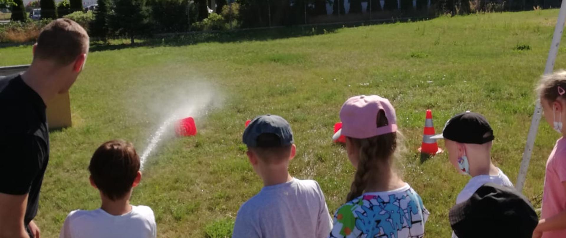 Dzieci stoją i obserwują jak strażak i chłopiec leją wodę na pachołki.