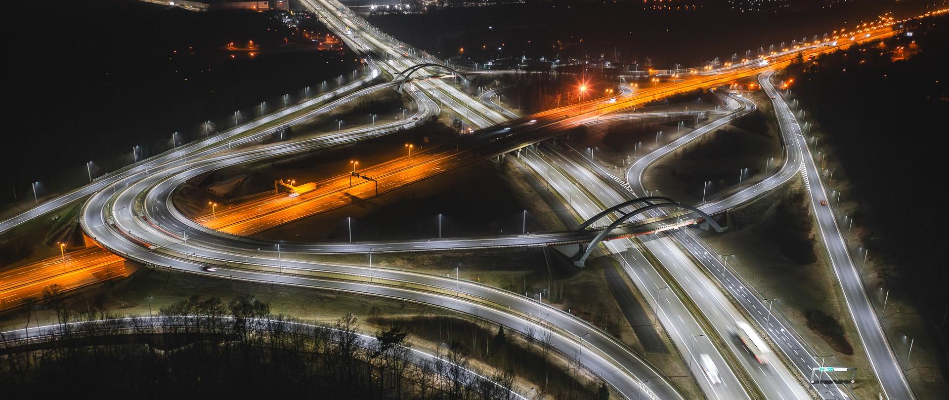Nocne zdjęcie lotnicze dużego węzła autostradowego. Jasne, białe światła oświetlają kręte zjazdy i estakady, tworząc skomplikowany układ dróg.