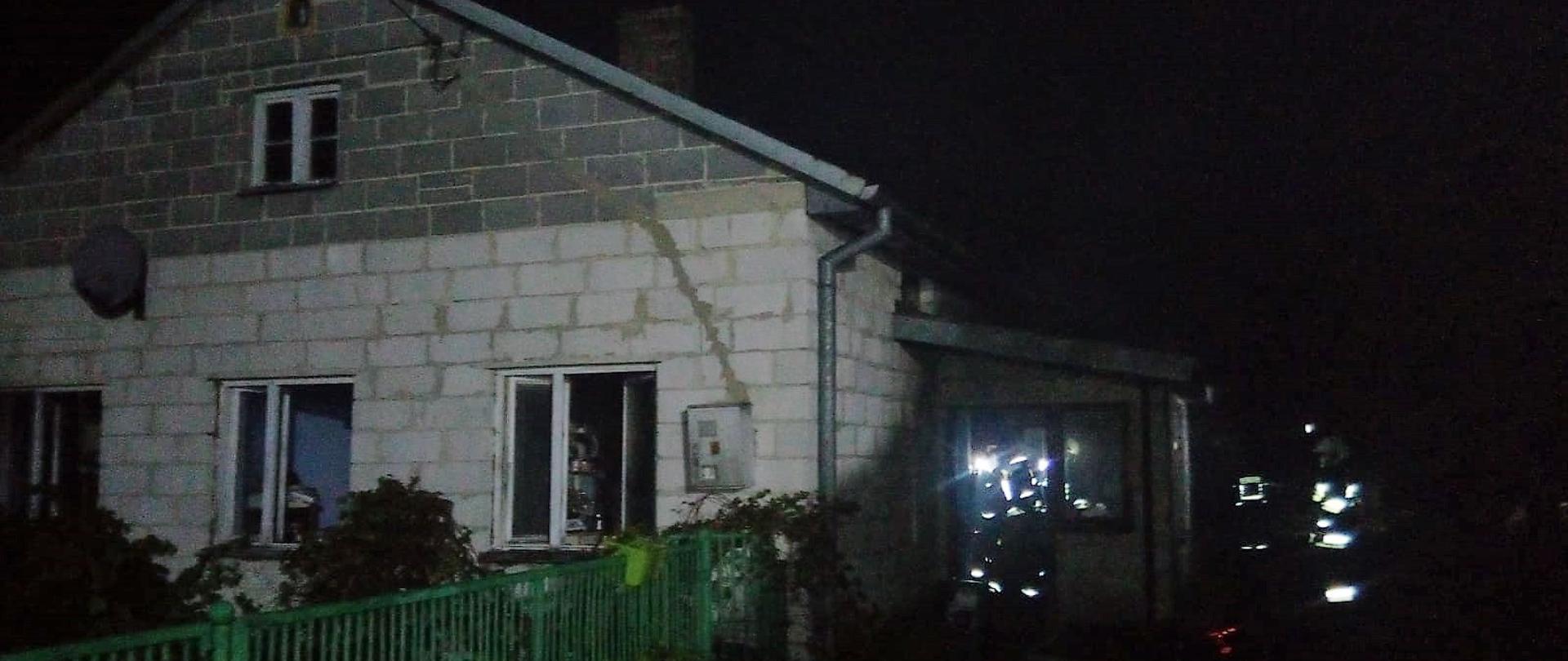 Zdjęcie przedstawia budynek jednorodzinny w którym doszło do pożaru. W ganku wejściowym widać postacie strażaków oraz węże gaśnicze rozwinięte na trawie.