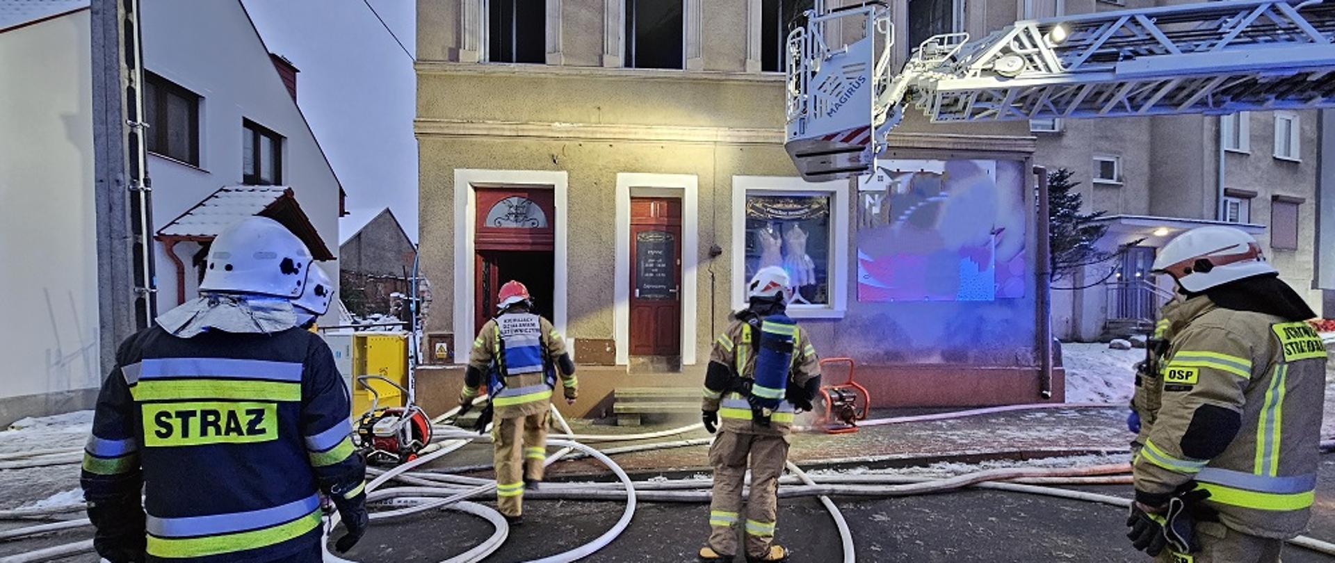 Zdjęcie przedstawia strażaków w trakcie pożaru kamienicy.
W tle budynek i samochody strażackie.
