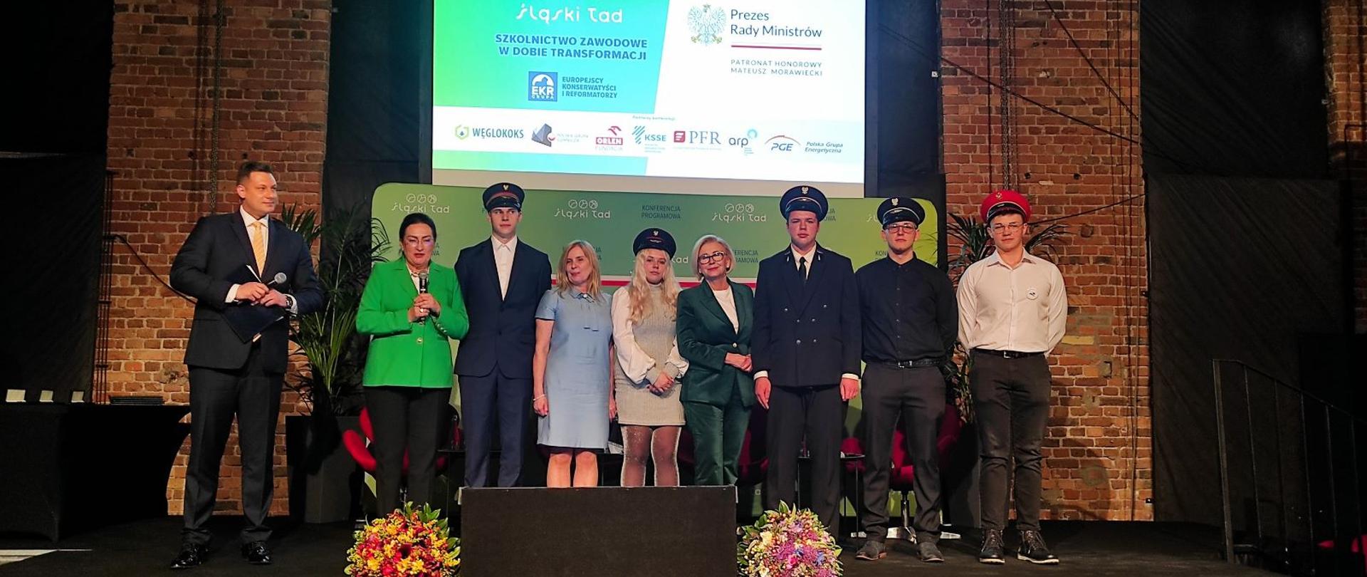 W sali z ceglanymi ścianami na podwyższeniu stoi 9 osób, za nimi zielona ścianka i duży ekran, przed nimi dwa bukiety kolorowych kwiatów.