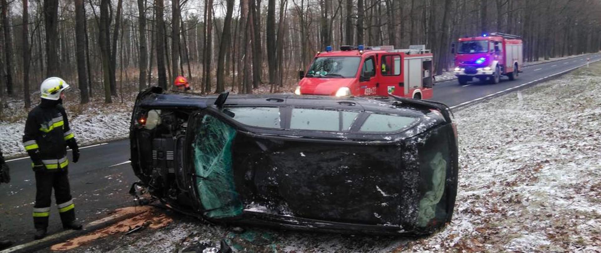 Droga krajowa numer dwanaście przy miejscowości Zajączek. Centralnie na zdjęciu znajduje się czarny samochód osobowy marki BMW, który znajduje się na poboczu w rowie. Samochód BMW położony jest na prawej boku pojazdu, jest to strona kierowcy. Samochód BMW w wyniku zderzenia z samochodem osobowym marki Alfa Romeo koziołkował. Widoczne uszkodzenia samochodu BMW to brak klapy przedziału silnika, zbita przednia szyba, uszkodzone boki i dach samochodu. Z lewej strony znajduje się strażak z jednostki ochotniczej straży pożarnej w białym hełmie i czarnym ubraniu specjalnym. za samochodem BMW w tle stoją na drodze krajowej czerwone samochody państwowej straży pożarnej i blokują przejazd drogi.