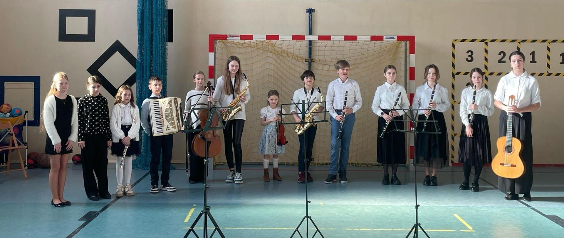 Uczniowie szkoły muzycznej stoją na sali gimnastycznej podczas audycji muzycznej w Koleczkowie. Prezentują swoje instrumenty pozując do zdjęcia.