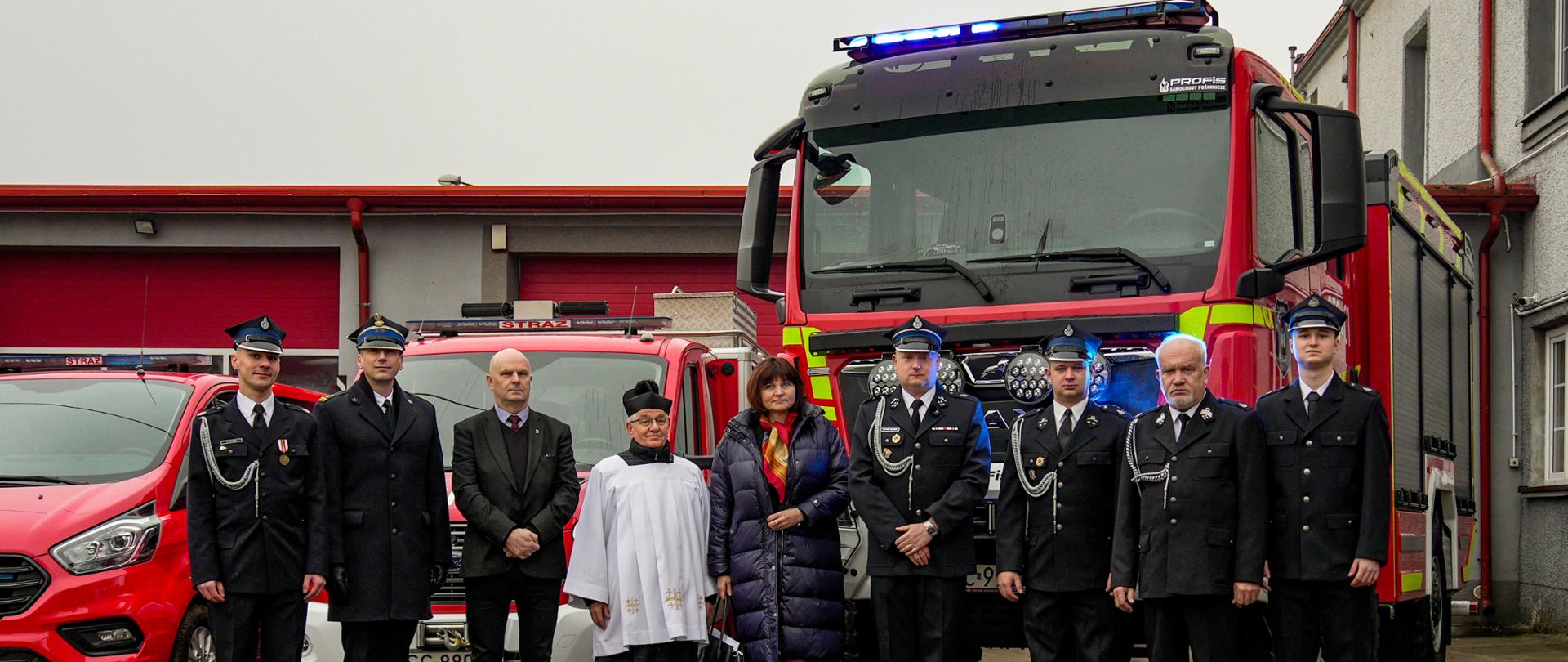 Na zdjęciu widzimy oficjalną, uroczystą fotografię wykonaną na terenie jednostki straży pożarnej. Na tle samochodów pożarniczych ustawione są w szeregu osoby, wśród nich strażacy w mundurach galowych, osoby cywilne i kapłan.