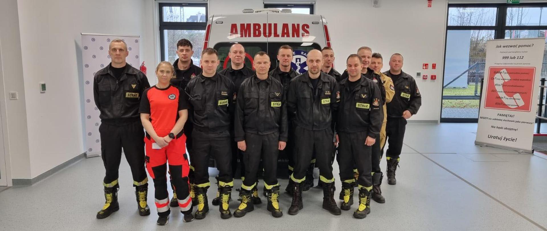 Strażacy stoją na tle symulatora karetki w centrum szkolenia wraz z instruktorem pierwszej pomocy.