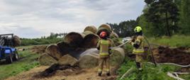 Pochmurny dzień na tle polany widać dwóch strażaków ubranych w piaskowe ubrania specjalne jeden ma czerwony hełm na głowie, drugi jaskrawo-żółty polewających wodą nadpalone baloty siana. Z lewego boku zdjęcia widać część niebieskiej ładowarki rolniczej. 