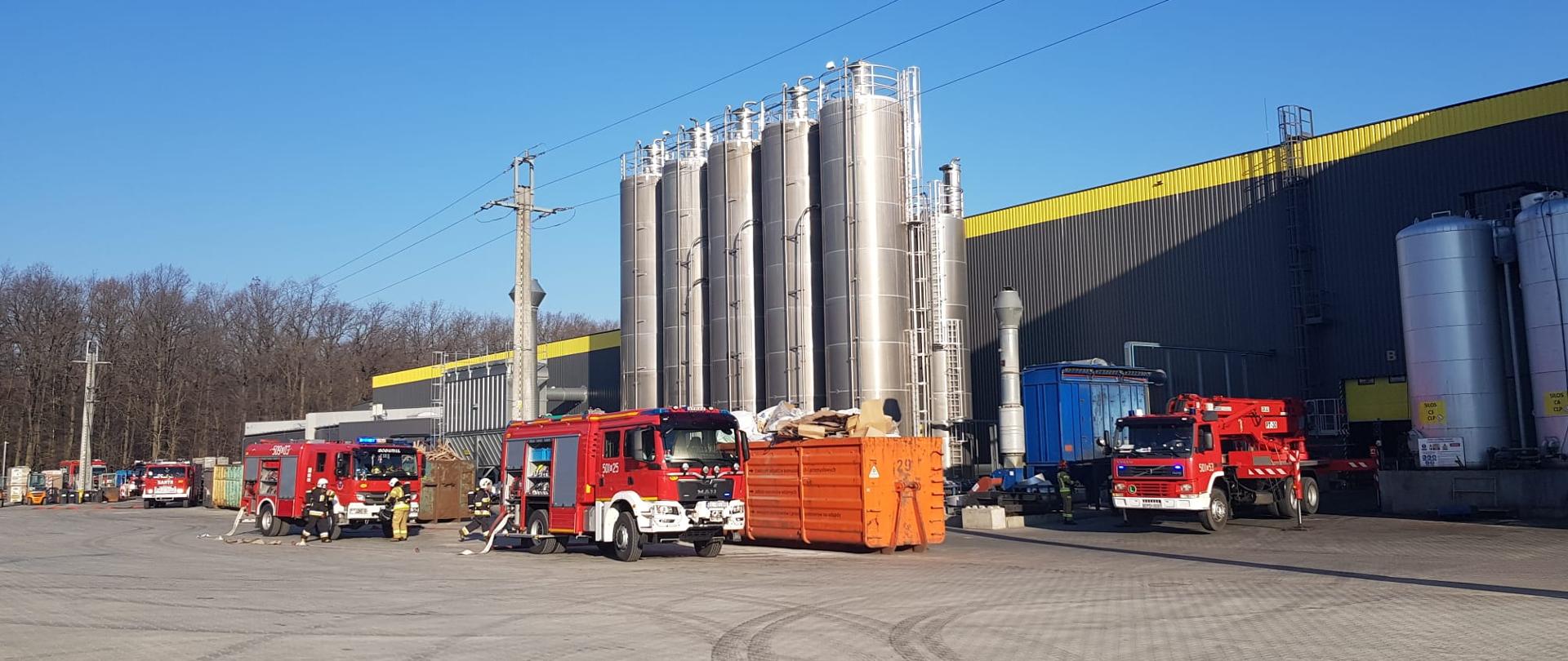 Trzy wozy strażackie stoją przed dużym zakładem. Przed samochodami strażacy rozwijają węże