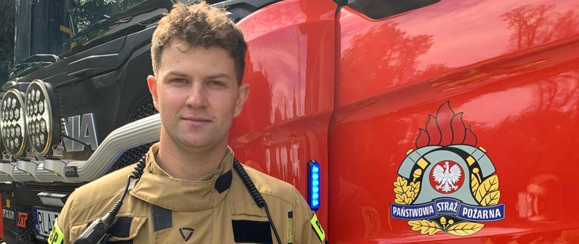 Zdjęcie przedstawia strażaka Konrada Skoczylasa w umundurowaniu specjalnym, trzymający w ręku hełm strażacki. W tle ciężki samochód ratowniczo-gaśniczy z napisem Łańcut na drzwiach kierowcy.