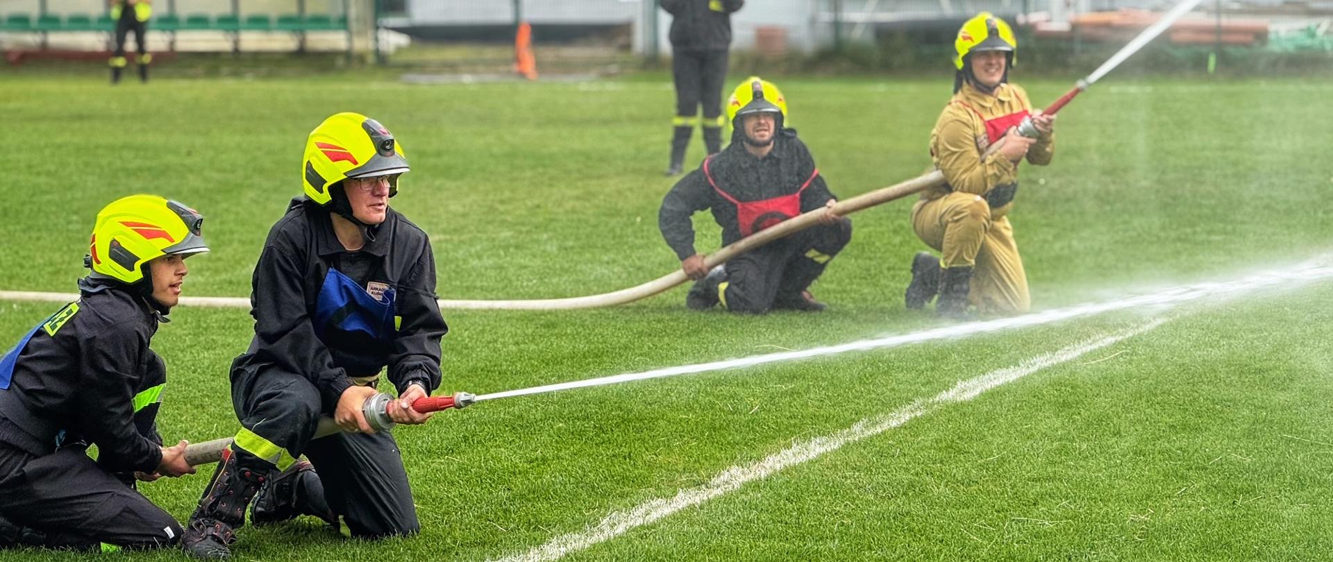 Zdjęcie przedstawia cztery osoby w mundurach strażackich, które klęczą na zielonym boisku. Dwie osoby na pierwszym planie kierują wężem, z którego tryska woda. W tle widać trybuny z zielonymi siedzeniami, a także inne osoby, które obserwują ćwiczenia