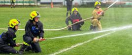 Zdjęcie przedstawia cztery osoby w mundurach strażackich, które klęczą na zielonym boisku. Dwie osoby na pierwszym planie kierują wężem, z którego tryska woda. W tle widać trybuny z zielonymi siedzeniami, a także inne osoby, które obserwują ćwiczenia