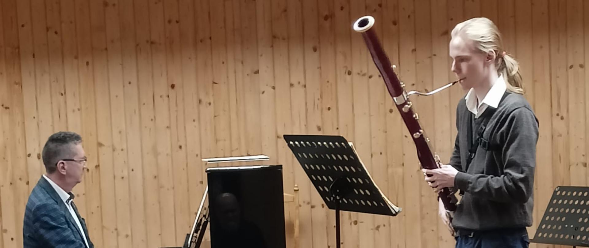 Nastolatek gra na fagocie, po lewej stronie mężczyzna gra pianinie.