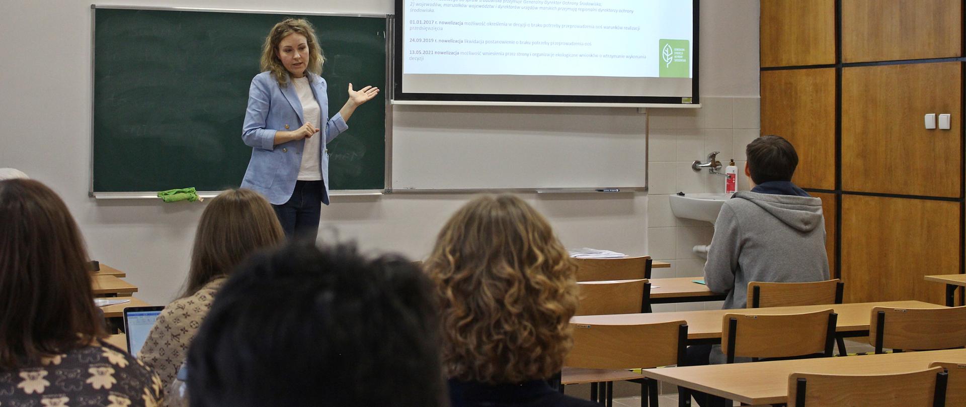 Sala wykładowa. Studenci zwróceni twarzami do tablicy. Na tle tablicy stoi młoda kobieta, która gestykuluje. W tle widoczna prezentacja. 