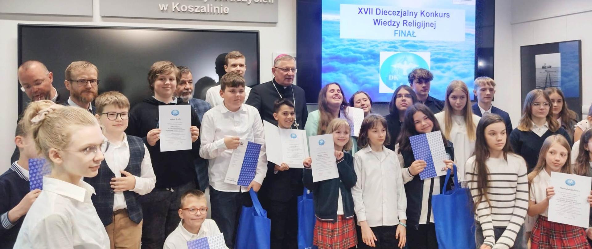 Uczniowie z dyplomami, w tle ksiądz i prezentacja multimedialna