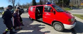 Widok z boku. Dwóch strażaków OSP w czarnych ubraniach koszarowych i maseczkach ochronnych pomaga starszej kobiecie w maseczce ochronnej wejść do czerwonego samochodu strażackiego. 
