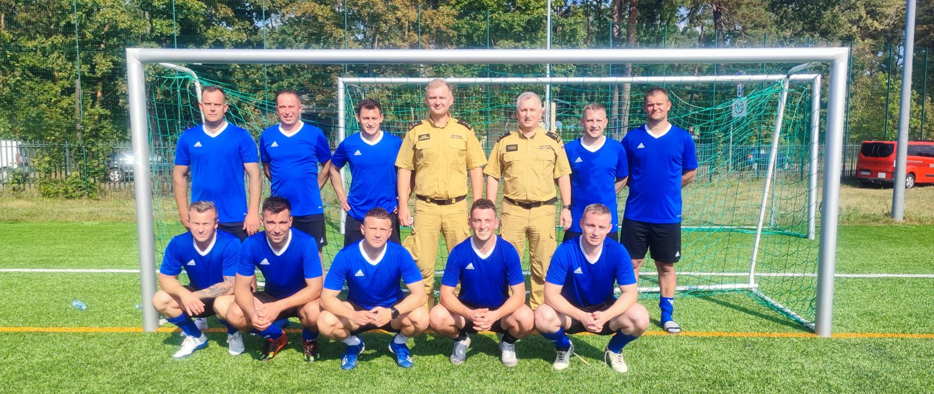 Na zdjęciu widoczna jest drużyna piłkarska w jednolitych strojach niebieskich, ustawiona na tle bramki. W środku stoi dwóch strażaków PSP ubranych w mundury służbowe. Za bramką widać samochody, w tym czerwony samochód strażacki oraz drzewa.