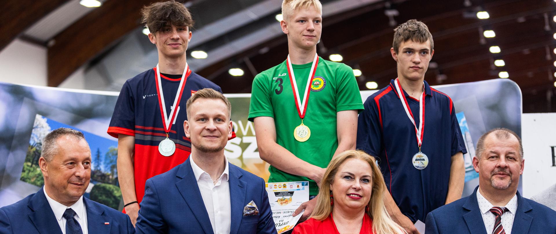 trzech chłopców stoi na podium, poniżej od lewej łódzki kurator oświaty, I wicewojeowa łódzki, kobieta w czerwonej sukience, starosta tomaszowski