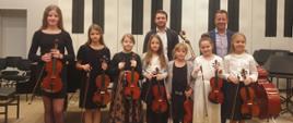 Siedem dziewczynek stoi w jednym rzędzie trzymając przed sobą skrzypce i smyczki, za nimi stoi dwóch mężczyzn.
