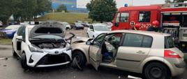Na zdjęciu widać dwa uszkodzone samochody po zdarzeniu oraz pojazd straży pożarnej w tle.