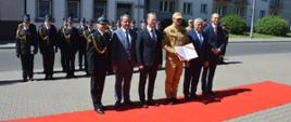 Plac przed komendą. Na czerwonym dywanie stoją przedstawiciele władz samorządowych oraz komendant powiatowy i dowódca JRG, który trzyma w rękach akt przekazania pojazdów pożarniczych. W tle pododdział funkcjonariuszy KP PSP w Rawiczu.