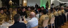 Uroczyste spotkanie w restauracji: przy długich stołach siedzą liczni strażacy Państwowej Straży Pożarnej w mundurach oraz zaproszeni goście; w tle nowoczesne wnętrze, choinka i osoba przemawiająca.