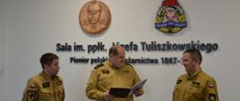 Strażak w umundurowaniu w kolorze musztardowym trzyma teczkę obok po obydwu stornach stoją funkcjonariusze Państwowej Straży Pożarnej na ścianie wisi logo Państwowej Straży Pożarnej oraz podpułkownika pożarnictwa pod nimi jest napis sala imieniem podpułkownika Józefa Tuliszkowskiego pionier polskiego pożarnictwa tysiąc osiemset sześćdziesiąt siedem myślnik. 