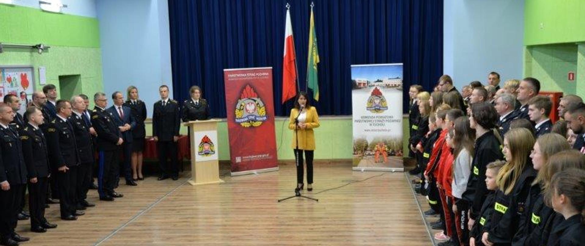 Zdjęcie przedstawia grupę osób. Na pierwszym planie stoi kobieta w żółtej marynarce przy mikrofonie. Po lewej stronie stoją goście oraz funkcjonariusze PSP w mundurach wyjściowych. Po prawej stronie stoją druhowie OSP oraz członkowie MDP. Strażacy mają na sobie mundury wyjściowe. Młodzież jest ubrana w czerwono-granatowy dres. Po prawej stronie znajduje się balkon. W tle widać niebieską kotarę, zielono-żółtą flagę Gminy Cekcyn, flagę Polski, dwie tablice z logiem PSP i mównicę z logiem PSP. Dookoła ściany są biało-zielone. Na podłodze są drewniane panele. 