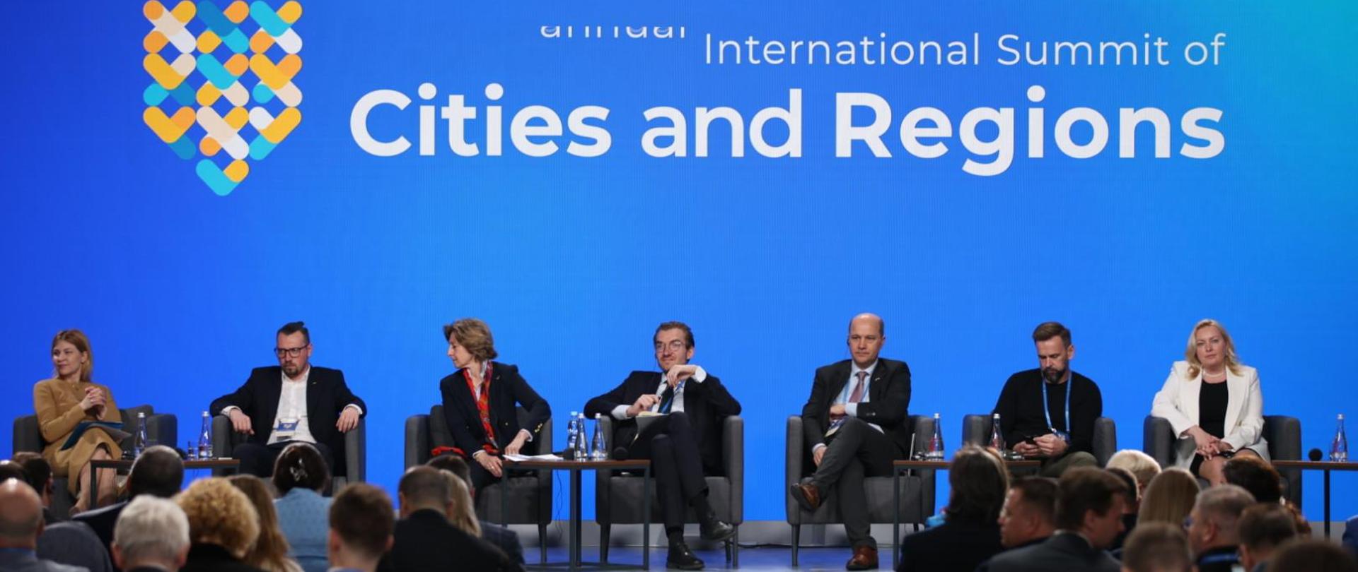 Grupa osób siedzi na scenie na sali.