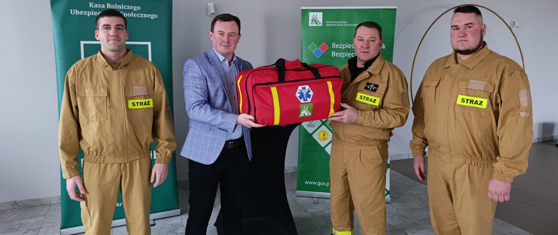 Czterech mężczyzn na tle dwóch zielonych banerów z logo KRUS. Dwóch z nich przekazuje sobie dużą, czerwoną torbę z logo KRUS i ratownictwa medycznego.