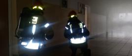 2 strażaków na kolanch przesuwa się po zadyminym korytażu, celm przeszukania pomieszczeń.