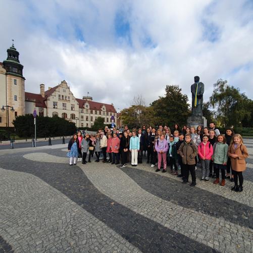 Uczniowie, rodzice i nauczyciele stojący na placu przed pomnikiem Adama Mickiewicza w Poznaniu.