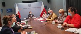 Przy podłużnym stole widoczni urzędnicy, Zachodniopomorski Komendant Wojewódzki Państwowej Straży Pożarnej st. bryg. Jarosław Tomczyk, Komendant Wojewódzki Policji. U szczytu stołu siedzi Wojewoda Zachodniopomorski Pan Zbigniew Bogucki. W tle widać baner wojewody oraz flagi Polski