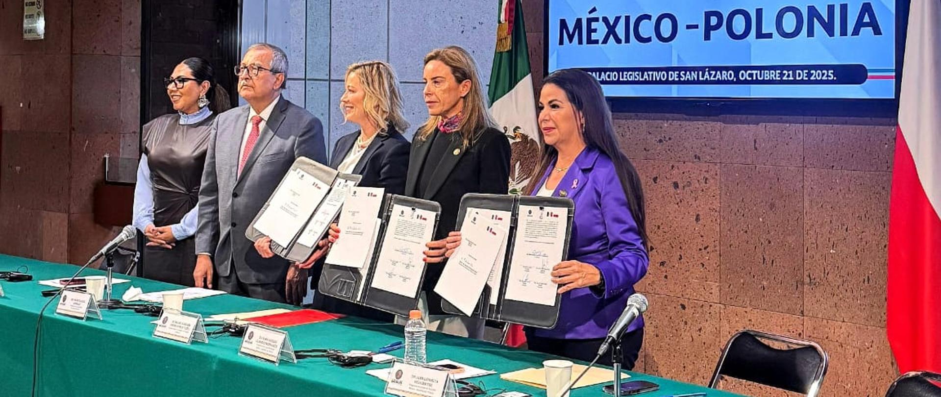 Na zdjęciu widać pięć osób stojących obok siebie w rzędzie, zwróconych przodem do obiektywu.
Od lewej do prawej stoją: posłanka Genoveva Huerta Villegas, poseł Pedro Vázquez González, Ambasador RP Agnieszka Frydrychowicz-Tekieli, posłanka María Angélica Granados Trespalacios oraz posłanka Alma Lidia De la Vega Sánchez.
Wszyscy są elegancko ubrani — w garnitury lub formalne stroje. Uczestnicy mają spokojne, skupione wyrazy twarzy, a niektórzy uśmiechają się lekko. W tle znajduje się sala parlamentarna z emblematami i flagami Meksyku, co nadaje scenie uroczysty i oficjalny charakter.
Zdjęcie przedstawia moment inauguracji Grupy Przyjaźni Meksyk–Polska.