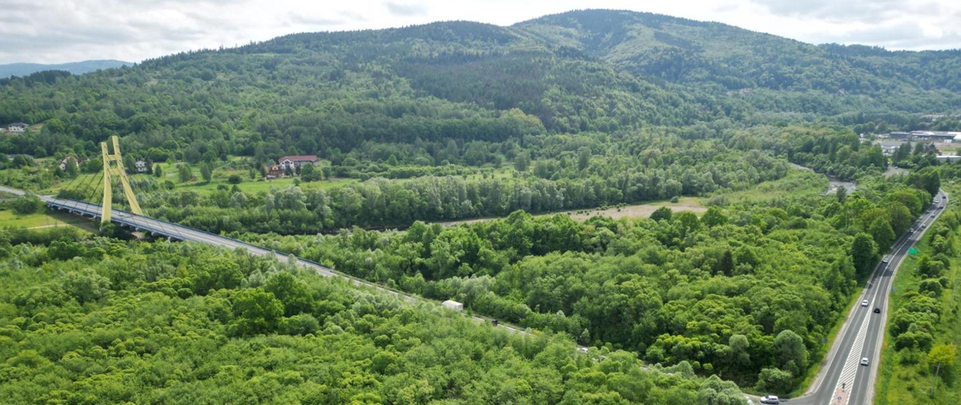 Widok z lotu ptaka na drogę i most w otoczeniu drzew. W tle widać niskie góry