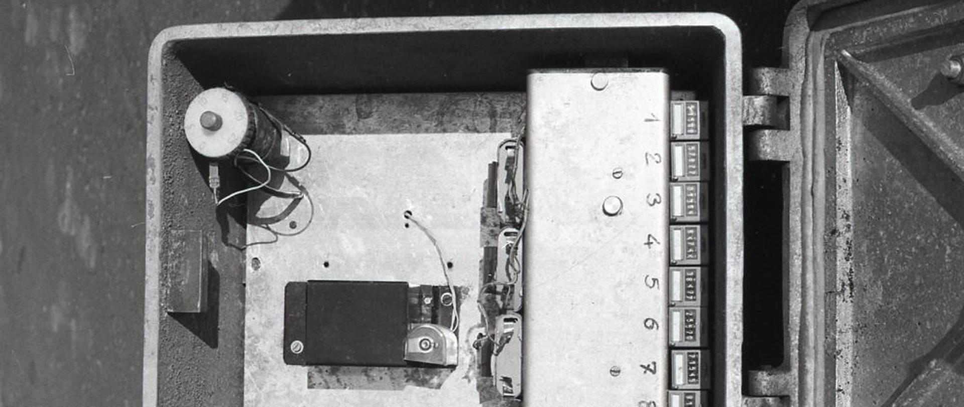 Widok na elementy przyrządu pomiarowego wewnątrz metalowej skrzyni. W środku osiem liczników analogowych, cewki elektryczne. 