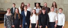Grupa uczniów stoi na scenie sali koncertowej, w tle widoczny jest fprtepian
