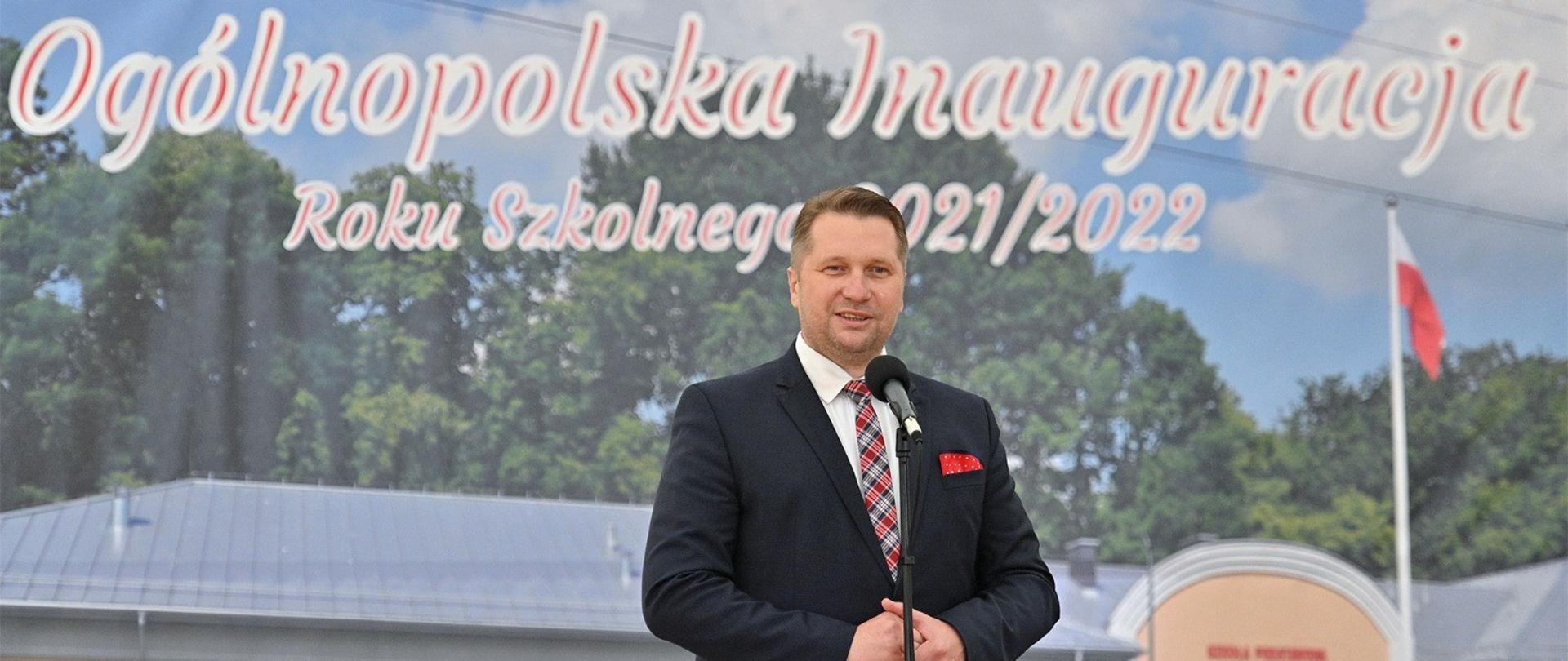 Minister Przemysław Czarnek podczas ogólnopolskiej inauguracji roku szkolnego 2021/2022 przemawia do mikrofonu. W tle zdjęcie nowo wybudowanej szkoły. 
