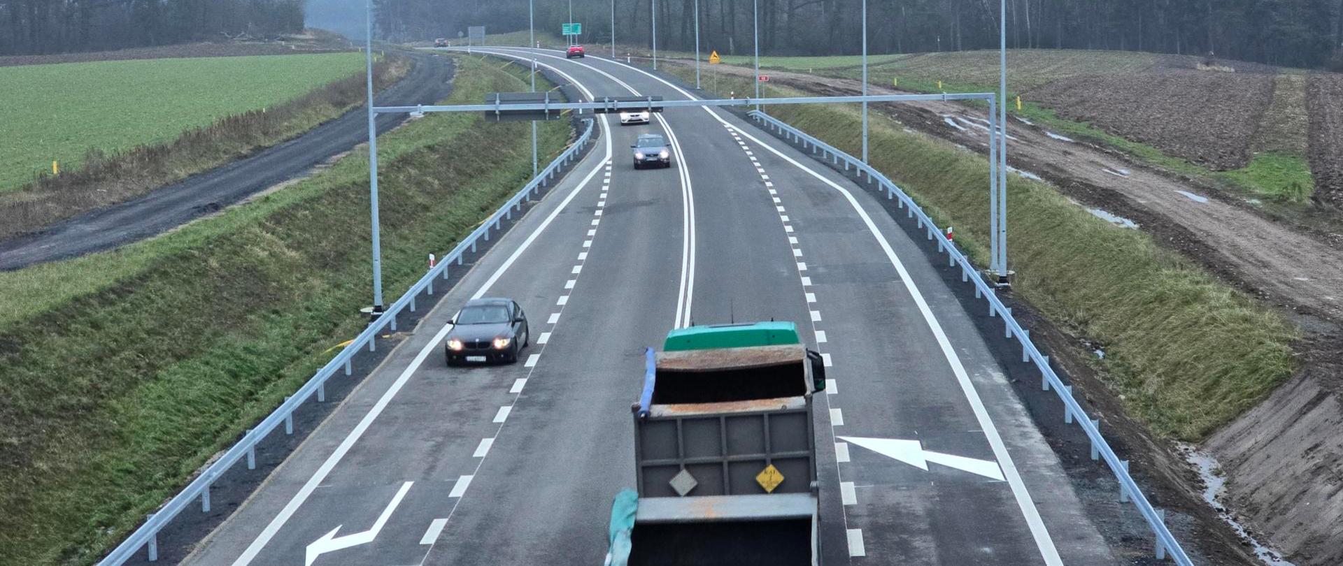 Widok na dwupasmową drogę z wyraźnymi białymi liniami i strzałkami kierunkowymi. Na jezdni porusza się kilka samochodów osobowych oraz ciężarówka z otwartą skrzynią ładunkową. Po obu stronach drogi znajdują się metalowe bariery ochronne, a w tle widoczne są pola uprawne i las. Niebo jest pochmurne, a krajobraz ma zimowy charakter.