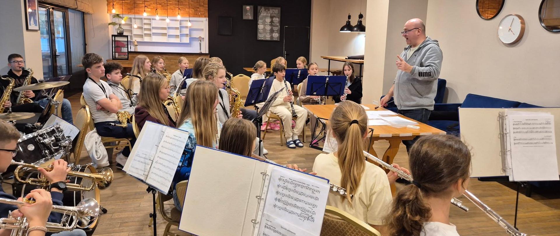 Próba orkiestry dętej podczas obozu kondycyjnego. Uczniowie siedzą i grają. Po lewej stronie dyrygent Tomasz Winczewski w sposób dynamiczny prowadzi próbę. 