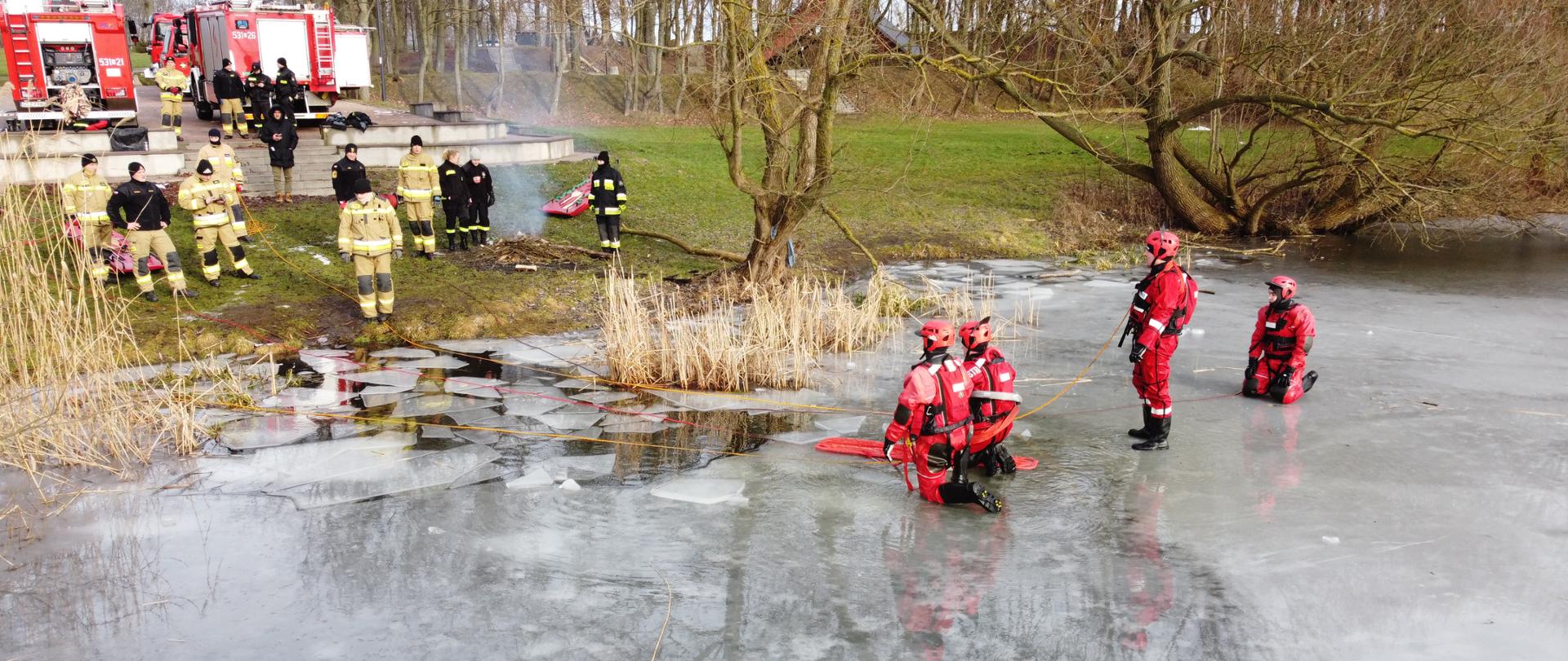 czterech ratowników w ubraniach do pracy w wodzie znajduje się na taflo lodu, pozostali ratownicy asekurują ich z brzegu