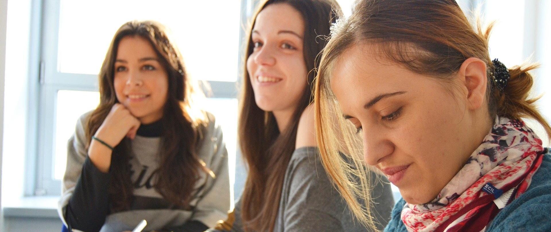 Trzy uczennice w wieku szkoły ponadpodstawowej siedzące w ławce szkolnej przy wypełnianiu ćwiczeń. 