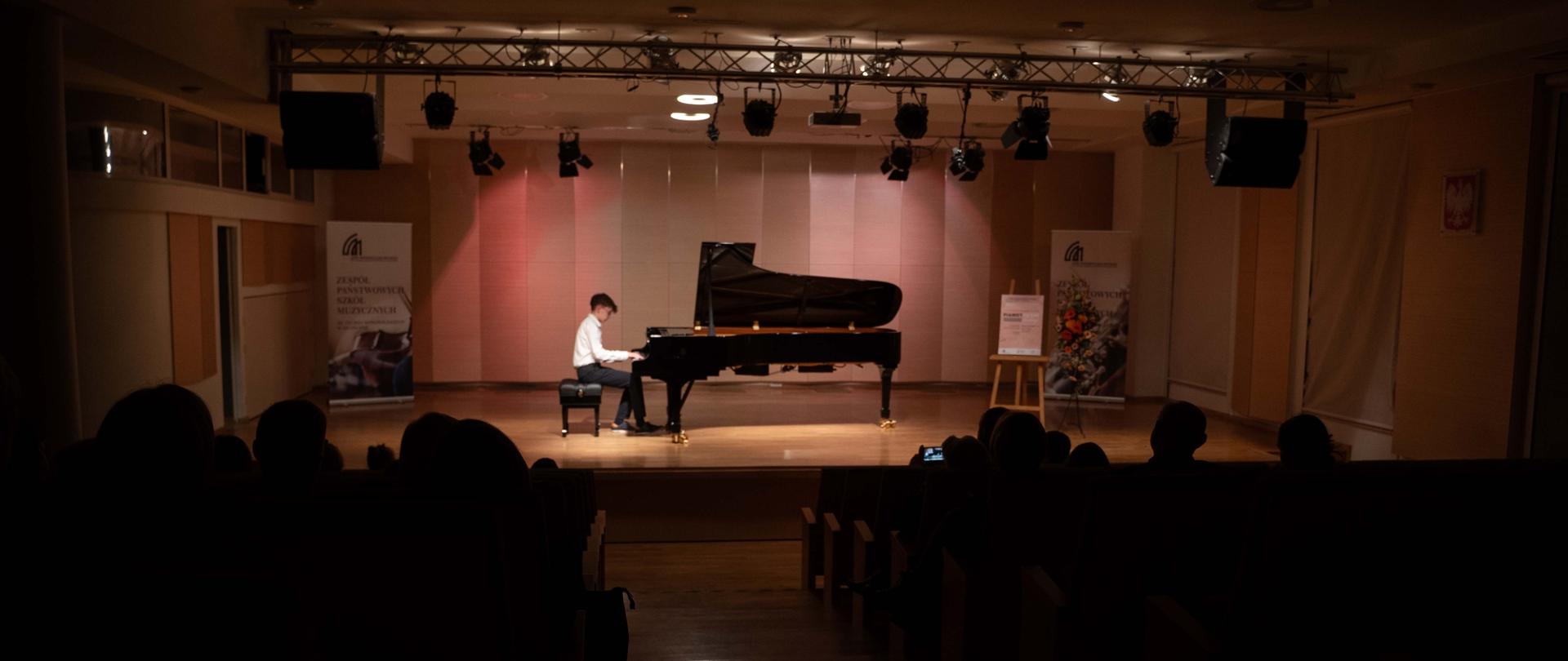 Zdjęcie przedstawia innego ucznia młodszej klasy grającego na fortepianie na scenie sali koncertowej, ujęcie z perspektywy tyłu widowni, widoczne banery szkoły, plakat koncertu oraz dekoracja kwiatowa
