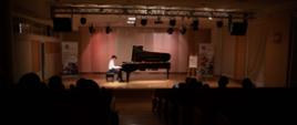 Zdjęcie przedstawia innego ucznia młodszej klasy grającego na fortepianie na scenie sali koncertowej, ujęcie z perspektywy tyłu widowni, widoczne banery szkoły, plakat koncertu oraz dekoracja kwiatowa