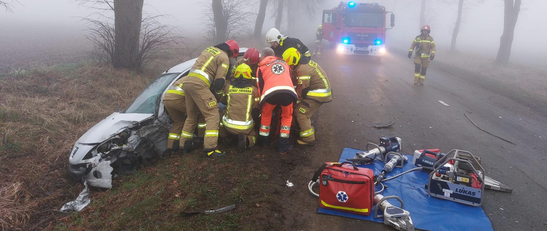 Strażacy i ratownik medyczny przy uszkodzonym białym samochodzie osobowym wyciągają poszkodowanego. Na jezdni rozłożony sprzęt hydrauliczny i torba medyczna. W tle widać mgłę i stojący samochód ratowniczo-gaśniczy.