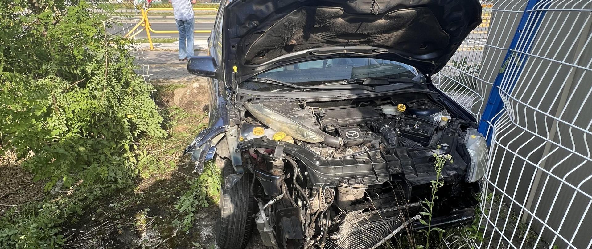 Zdjęcie przedstawia zniszczony samochód osobowy. Po prawej stronie widać fragment uszkodzonego ogrodzenia posesji, a za autem zniszczone, żółte barierki zabezpieczające ścieżkę rowerową. 