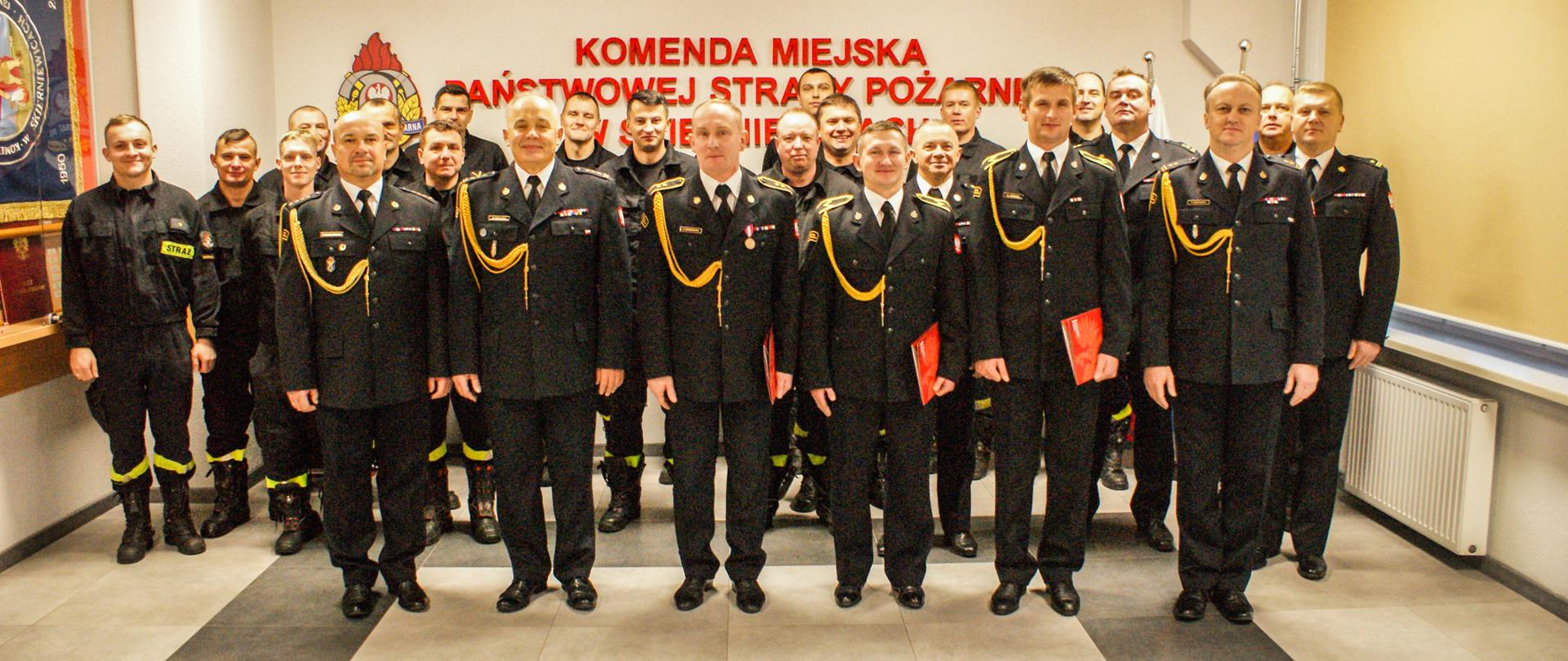 Zdjęcie przedstawia grupę strażaków w mundurach wyjściowych koloru czarnego na tle jasnej ściany z napisem z godłem państwowym i logo PSP