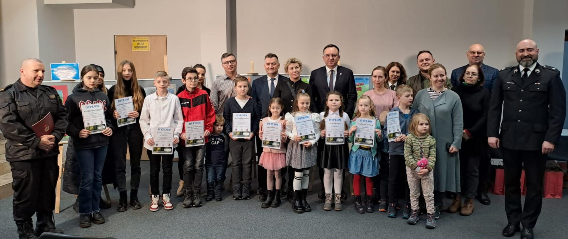 Na dużej sali na tle białej ściany stoją obok siebie dzieci z wyróżnione w konkursie z dyplomami oraz organizatorzy konkursu.