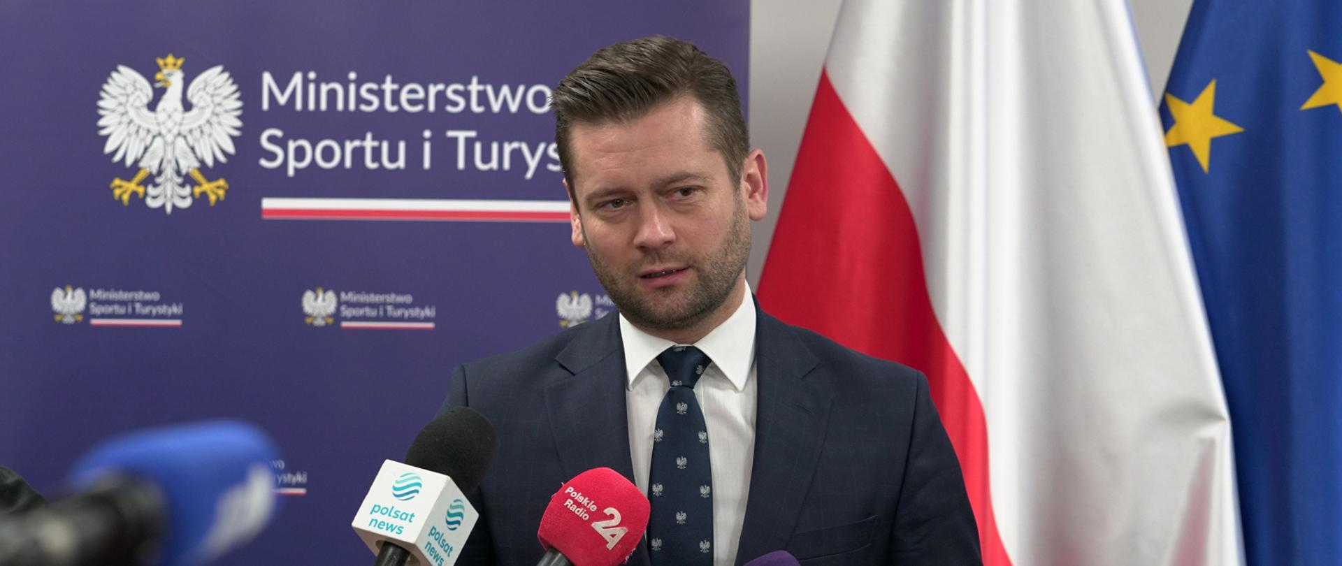 Na zdjęciu znajduje się elegancko ubrany mężczyzna, który jest w trakcie wystąpienia publicznego. W tle znajdują się flagi Polski i Unii Europejskiej oraz granatowa ścianka wizerunkowa Ministerstwa Sportu i Turystyki, na której znajduje się godło Polski.